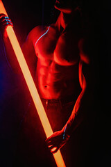 Muscular strong guy with naked torso abs holds neon light tube in dark room