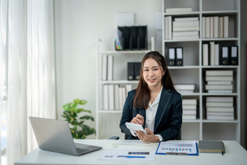 A woman in a business suit sits at a desk with a laptop and tablet. She is smiling and she is working on a project