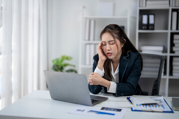 A woman is sitting at a desk with a laptop and a stack of papers. She is looking at the laptop and she is in a state of distress