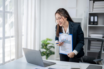 A woman in a business suit is standing in front of a laptop computer and holding a clipboard. She is excited or happy about something, possibly related to her work