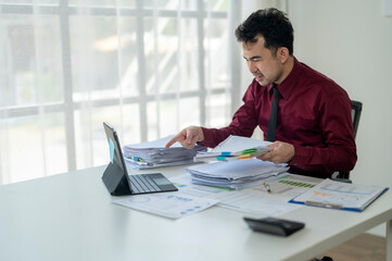 A man in a red shirt is sitting at a desk with a laptop and several papers