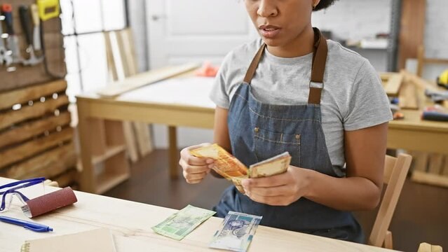 A focused woman in an apron counts south african rand bills at a carpentry workshop table surrounded by tools.