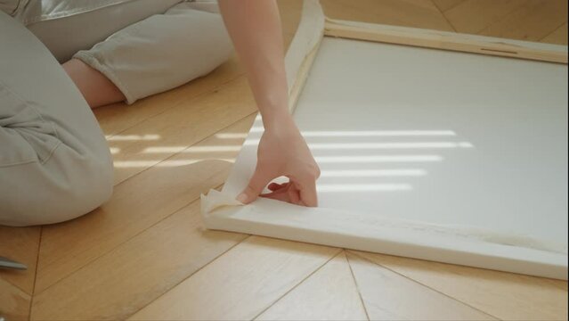 Close-up of young woman's hands stretching canvas on stretcher, fixing fabric to wooden frame with construction stapler. Pleasant pastime for hobby on sunny weekend day at home. Creativity diy