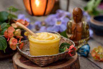 Ghee butter in a glass container on a wooden table, with flowers, a candle lantern, and a buddha statue in the blurred background.