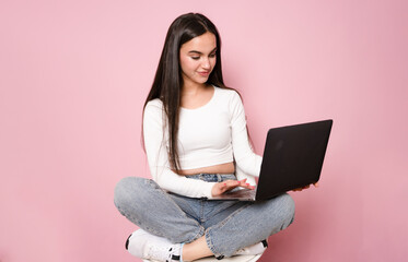 Happy young woman sitting on a chair with crossed legs and using laptop on pink background.