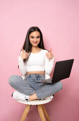 Happy young woman sitting on a chair with crossed legs and using laptop on pink background.