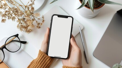 Top view of female hands holding smartphone with blank screen on white office table with plants glasses paper, flat lay with space for text