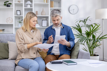 Obraz premium Middle-aged couple reviewing financial documents at home on the couch, looking concerned. Concept of financial planning, budgeting, and dealing with paperwork.