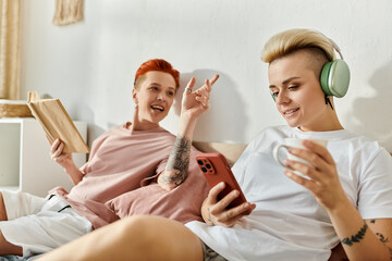 A lesbian couple with short hair sits on a bed, wearing headphones as they enjoy music together in their bedroom.