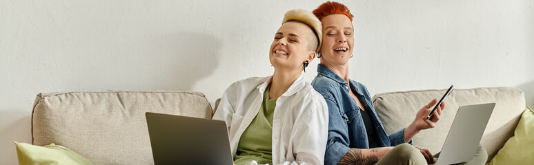 Lesbian couple with short hair engrossed in work on laptops while sitting closely on a couch at...