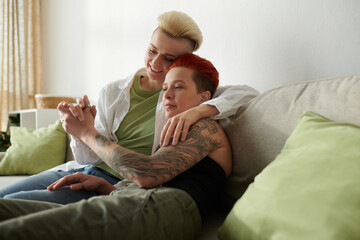 Two lgbt women with tattoos sit comfortably on a couch, sharing a moment of togetherness and...