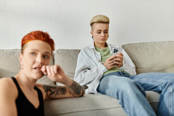 Two people, a lesbian couple with short hair, sit on a couch absorbed in phone, disconnected from...