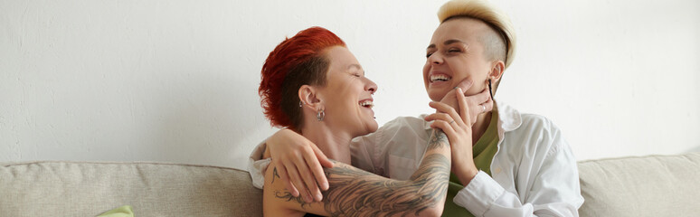 Two women with short hair share a warm hug on a cozy couch, expressing love, comfort, and...