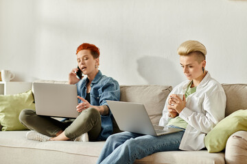 Two women with short hair sit side by side on a couch, each absorbed in their own laptop screen.