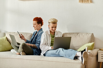 Two women with short hair sitting on a couch, engrossed in their laptops in a cozy home setting.