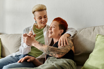Two women, a lesbian couple, with short hair and tattoos, relax together on a couch.
