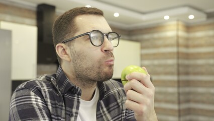 Young man in glasses sitting at the kitchen and eating green apple. Slow motion