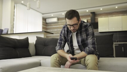 Sad man in glasses reading message on the phone, sitting on the sofa at home