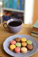 Purple plate filled with pastel macarons, cup of tea or coffee, vintage books and reading glasses on the table. Colorful bookcase in the background. Selective focus.