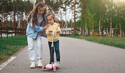 Obraz premium Family care concept. A little girl teaches her sister to ride a scooter in the park.