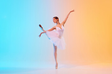 Obraz premium Young ballerina in elegant attire, tutu and pointe, performing in neon light against blue-orange gradient background. Graceful. Concept of art, movement, classical and modern fusion, beauty, fashion.