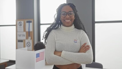 A confident woman with a 'i voted' sticker stands with crossed arms in a polling station with us flags.