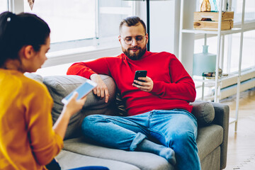 Young male and female sitting on sofa at home interior ignoring talking to each other spending time on modern smartphones, couple addicted to online communication and apps using mobile phones