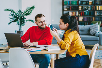 Fototapeta premium Positive couple using technology and wireless connection at home interior for freelance, woman using mobile phone for making booking online talking with her positive boyfriend working on laptop
