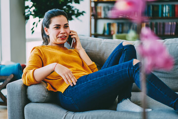 Smiling caucasian woman making telephone call lying on comfortable couch in living room, positive...