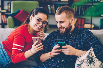 Cheerful romantic couple watching funny video from social networks on mobile phone resting on sofa,...