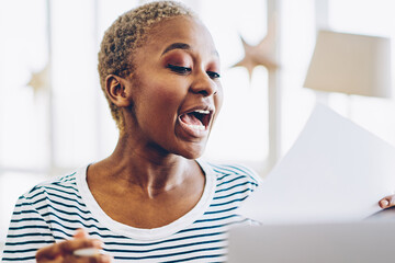 Overjoyed african american hipster girl excited with good news reading from paper sitting near...