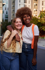 Vertical image of two multiracial women embraced outdoors in the city street, smiling while looking away.