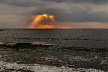 Rays of golden sunlight shoot through the clouds at sunrise over the Mediterranean Sea.