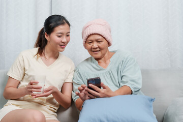 Young woman and elderly woman sitting together, looking at smartphone. Elderly wearing pink beanie, indicating cancer treatment. Both smiling, emotional support and connection. Warm, caring atmosphere