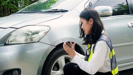 Woman in safety vest examines car with phone, concerned, foliage in soft focus behind. Concept of...
