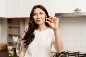 Confident Young Woman In Kitchen Showing Omega-3, Modern Interior Background. Concept Of Satisfaction, Approval, Good Service. Natural Light, Casual Style.
