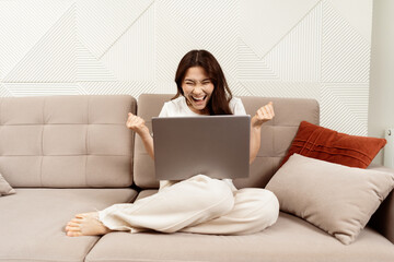 Excited Young Woman Celebrating Success On Laptop At Home, Sitting On Sofa, Expressing Joy And Happiness, Modern Interior