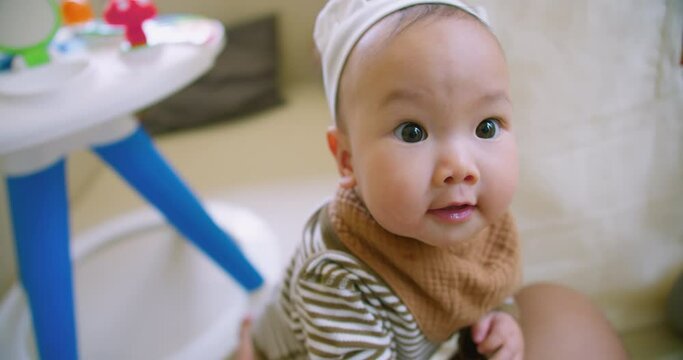 Adorable baby with wide eyes wearing a cute headband and a brown bib looking up curiously at the camera in a bright indoor