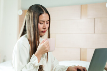 Young Asian woman drinking from pink mug while using laptop in bedroom. Wearing white blouse, long hair. Thoughtful expression, working from home. Comfortable, modern bedroom setting.