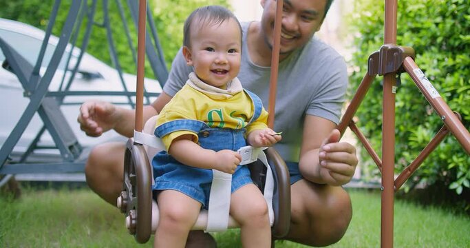 A joyful moment of a father pushing his smiling baby in a swing at a garden playground. The baby, dressed in a yellow shirt and denim overalls, is safely strapped in, enjoying the outdoor playtime.