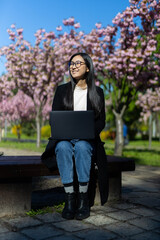 A young Japanese student works on her laptop in the blooming sakura park, enjoying the outdoors and studying.