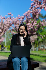 A successful Korean woman uses technology in the park, enjoying the outdoors while staying...