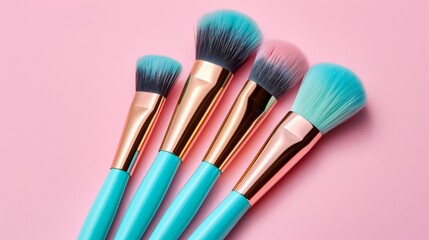A set of four makeup brushes with turquoise handles and pink and blue bristles, arranged on a pink background