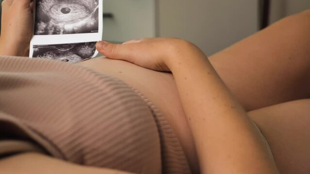 Girl in pink top gently running her hand over her pregnant belly. In the other hand she holds an ultrasound image with the stage of the baby's development. Anticipation and joy of motherhood