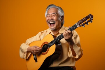 Portrait of a content asian man in his 80s playing the guitar while standing against soft green...