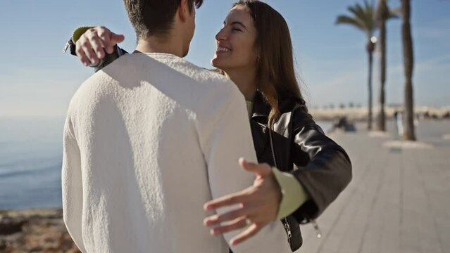 A couple embraces joyfully on a sunny beach promenade with palm trees, featuring a woman in a leather jacket and a man in a white sweater.