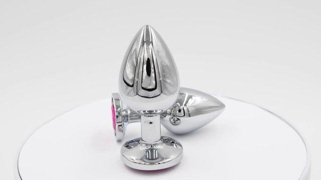 Close-up side view of two metallic butt plugs with red round crystals spinning on white background. Soft focus. Real time video. Adult sex toys theme.