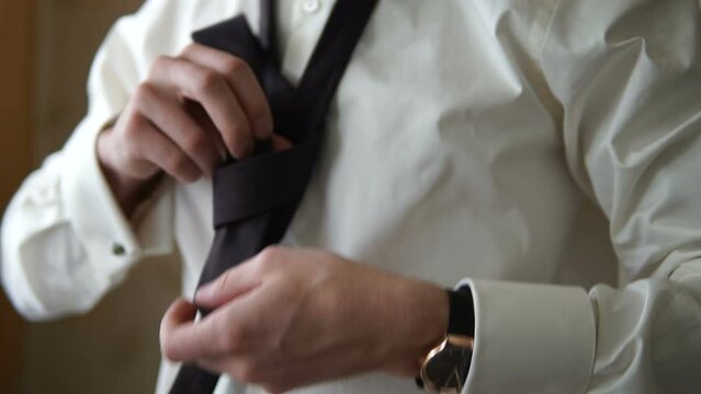 A man in a white shirt with a watch and cufflinks ties a knot on his tie. A manager is getting ready for work. A boss is putting on a tie. A businessman is getting ready to meet a partner. Getting rea