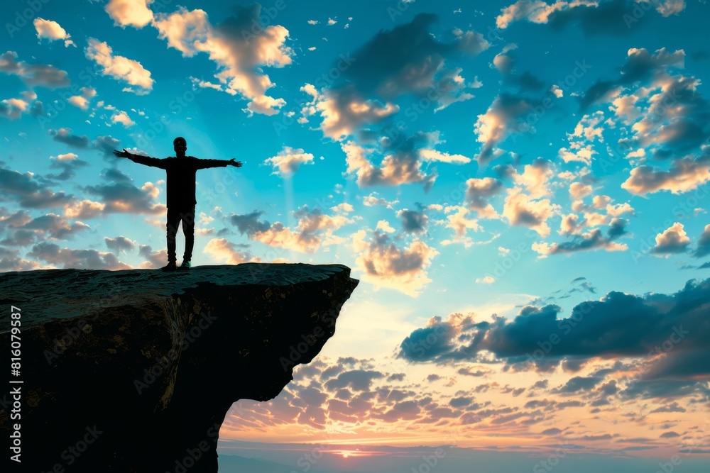 Wall mural man in silhouette stands on the edge of cliff with open hands against blue sky with clouds at sunris