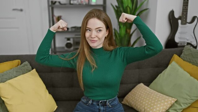 Smiling proudly, young blonde woman flexes arm muscles at home. sporting a sweater, she embodies a fit lifestyle and fitness concept. healthy, powerful, confident!